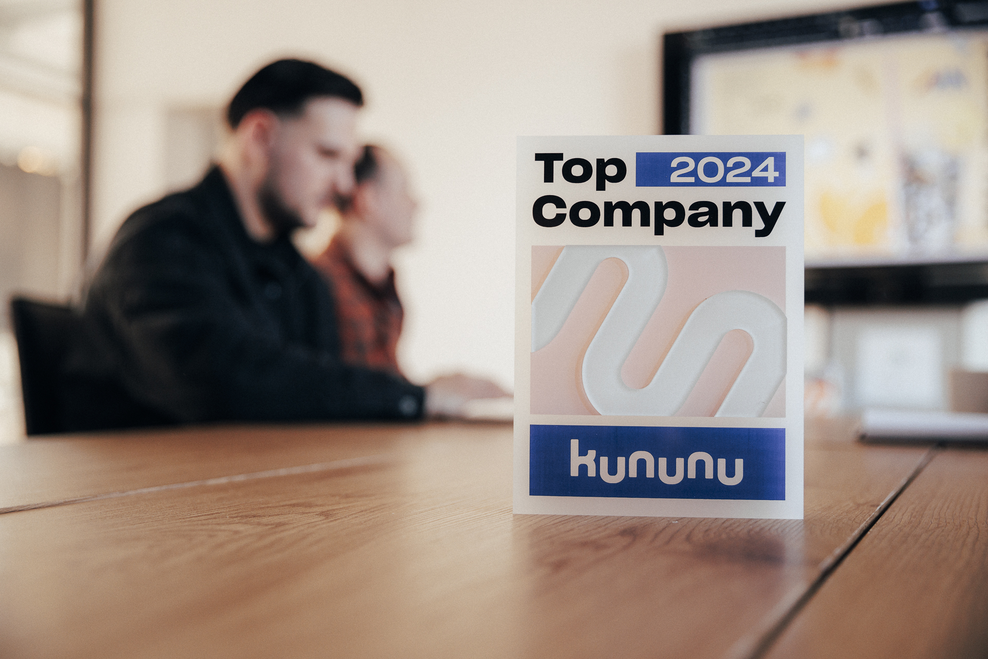 Im Hintergrund zwei Personen an einem Tisch, im Vordergrund eine Auszeichnung mit dem Namen "Top 2024 Company" von kununu