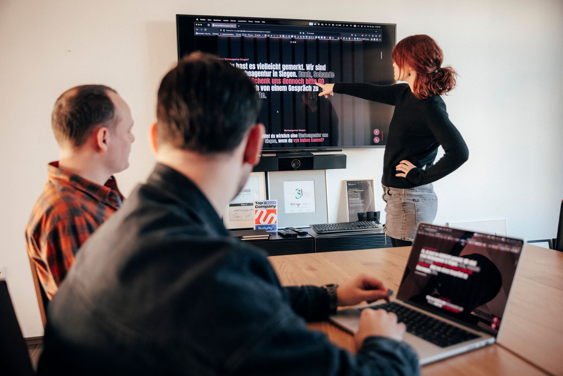 Zwei Personen sitzen an einem Tisch vor einem Laptop, während eine weitere etwas an einem Bildschirm zeigt