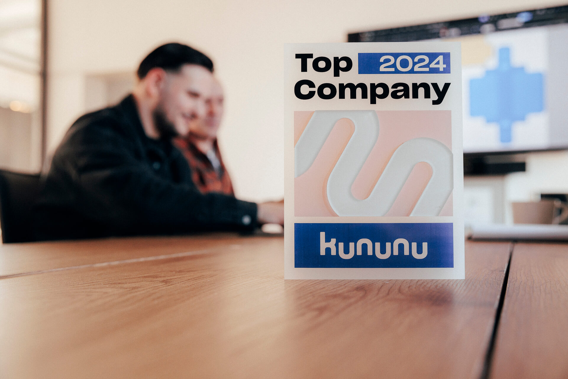 Im Hintergrund sitzen zwei Personen an einem Tisch, im Vordergrund steht eine Auszeichnung für "Bester Arbeitgeber"