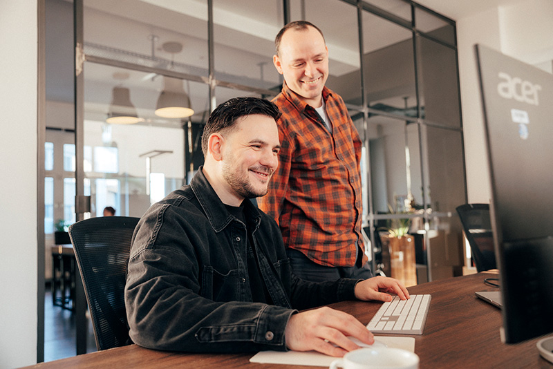 Zwei Personen stehen und sitzen lächelnd vor einem Computer